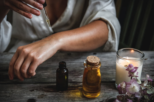 Woman applying oils to the skin of her arm using dropper. Oils, candle and flowers on wooden table